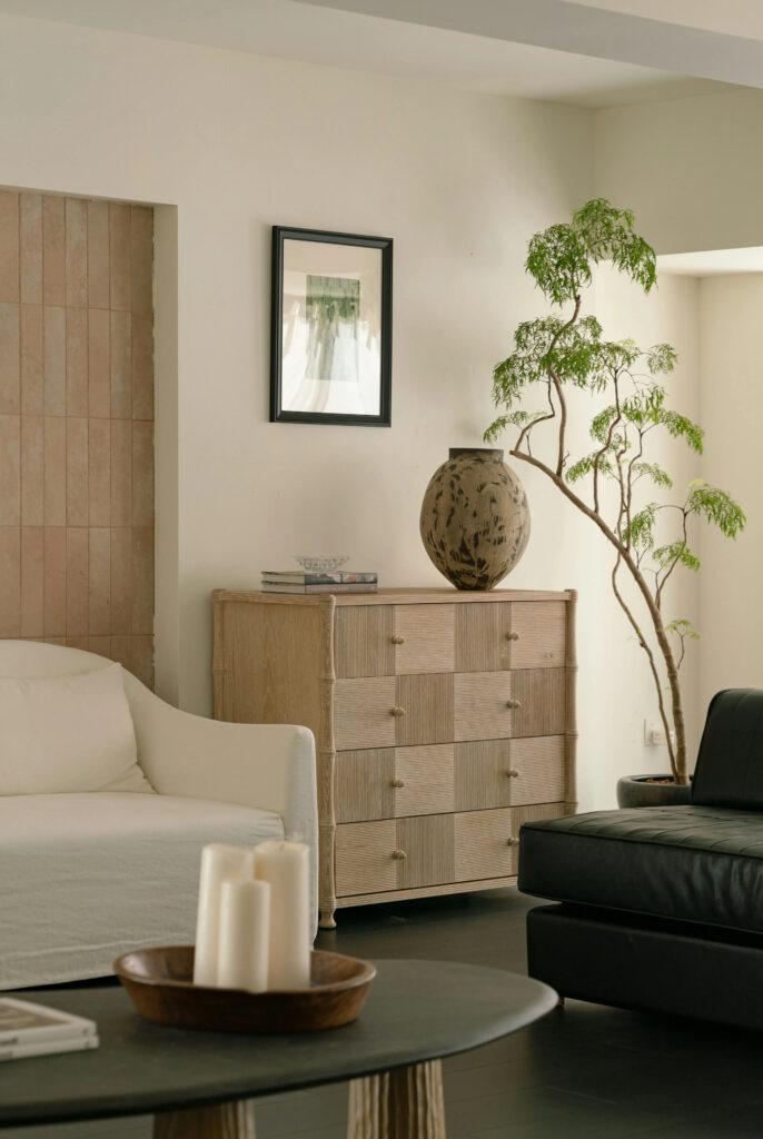 Stonowany salon z białą sofą, czarną leżanką i komodą z jasnego drewna, ozdobiony drzewkiem bonsai oraz misą świec.